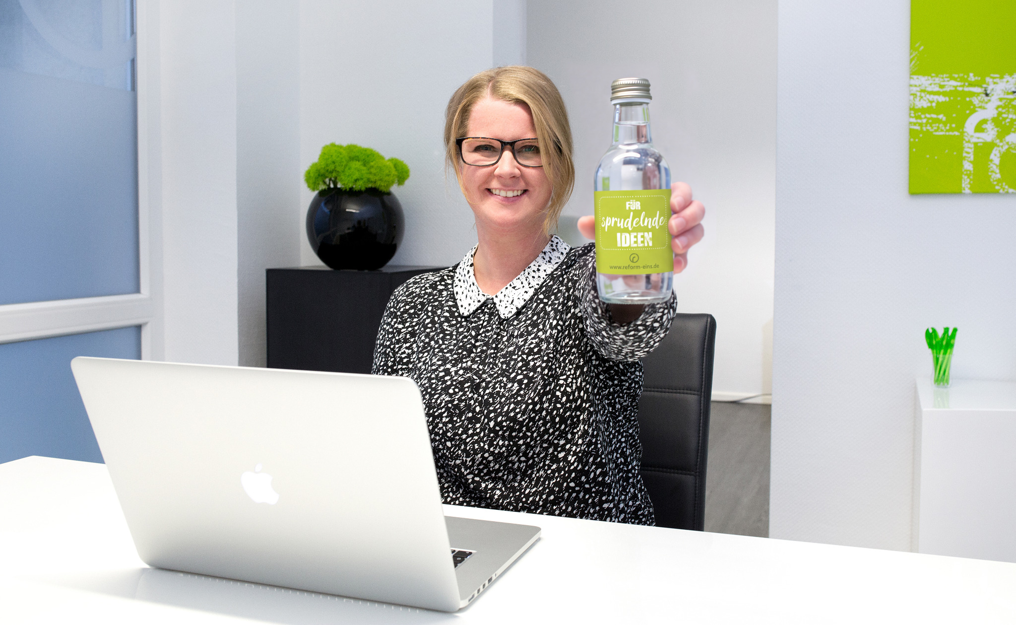 Stephanie Renelt lächelt im Büro und hält eine Flasche mit der Aufschrift „Für sprudelnde Ideen“.